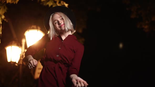 Millennial Hipster Woman with Blond Hairstyle Dancing Under the Lantern of City Street at Night. Hat