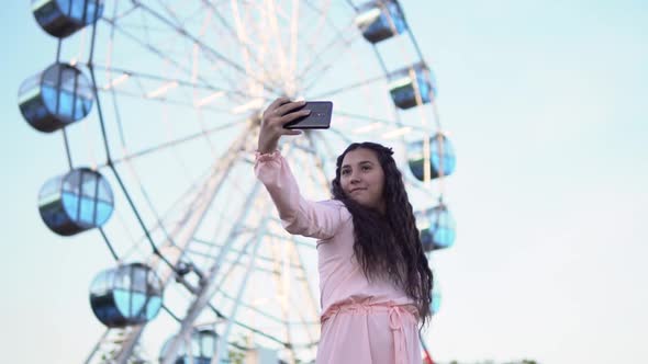 a Girl with Long Hair in a Dress Makes Selfie Using a Smartphone Standing Near the Ferris Wheel