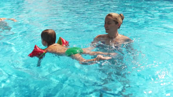 Mom in Bikini Plays with Baby in Oversleeves and His Sister in a Pool of Clear Water