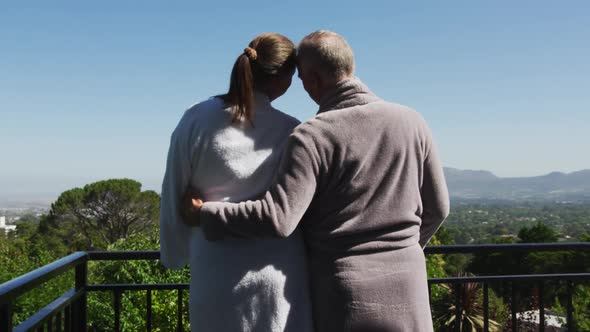 Rear view of caucasian senior couple in bathrobes embracing each other in the balcony at home