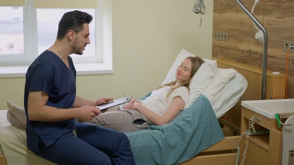 Side View Portrait of Smiling Positive Middle Eastern Male Doctor Sitting on Hospital Bed Talking to