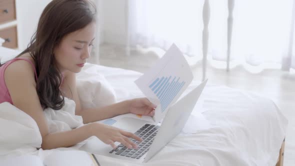 Beautiful freelance asian woman working and typing with laptop computer on bed in the bedroom.