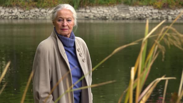 An Elderly Woman Stand By a Lake and Looks Solemnly Into Distance