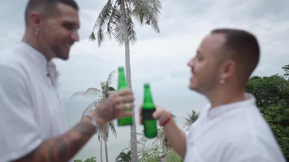 Male Gay Couple Clinking Bottles of Beer and Smiling While Resting on Tropical Island Standing at