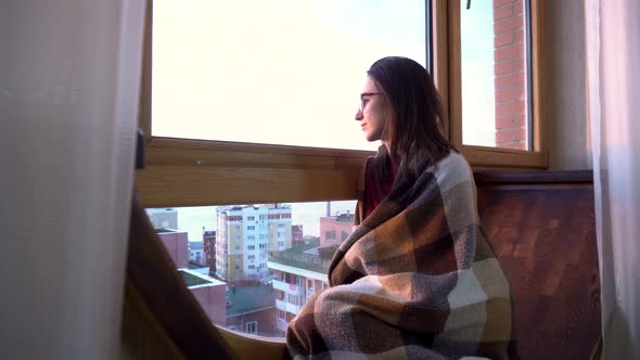 A Young Woman Is Sitting By the Window. A Woman Sits on a Windowsill Wrapped in a Plaid