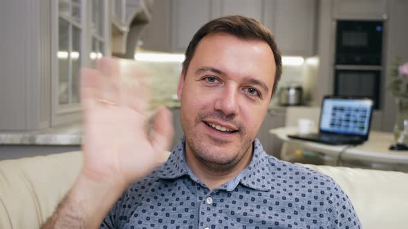 Young Adult Male Sitting on Sofa at Home Looks at Camera and Greets Colleagues or Students Family