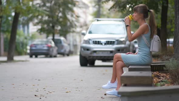 Young Woman Drinking Coffee From Paper Cup Sitting on City Street Bench in Summer Park