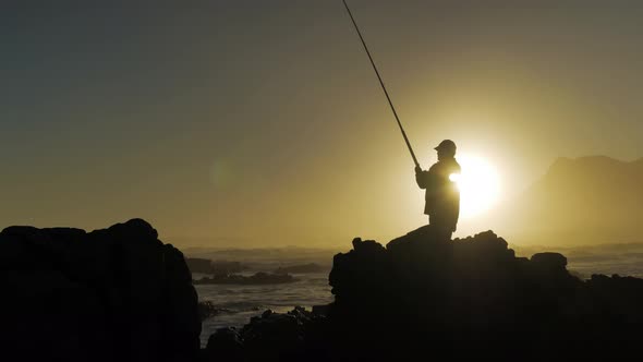 Fisherman on rocks reeling in fish during  golden sunset over ocean, silhouette against sky.