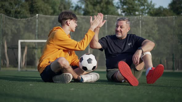 Positive Mature Father Amputee and Teenage Son Gesturing Highfive Sitting on Sunny Summer Stadium