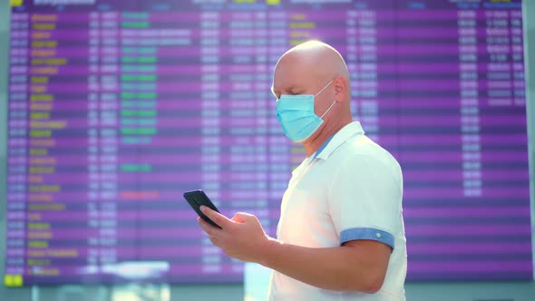 Male Air Passenger in Mask, Standing Against Departure Board at Airport. He Is Checking Flight