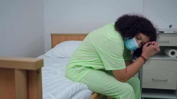 Exhausted Desperate Doctor in Covid19 Face Mask Sitting on Bed in Hospital Ward Praying