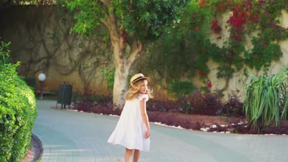 Little Girl in a White Dress and a Straw Hat is Merrily Strolling Along the Path