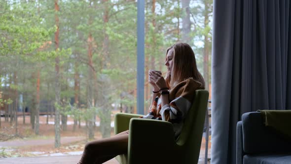 charming woman drinks tea by a large window overlooking a pine forest