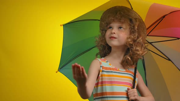 Curly Blond Girl in a Straw Hat with a Rainbow Umbrella is Checking for the Rain