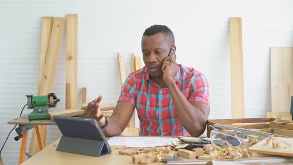 African american male carpenter speaking by phone with client  and making notes in woodworking shop