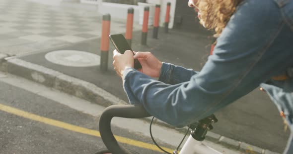 Happy biracial woman in city, sitting on bike using smartphone