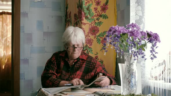 Portrait of an Old Woman in Glasses Reading a Newspaper While Sitting at a Table at Home