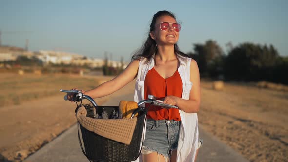 Medium Shot Portrait of Happy Confident Woman in Sunglasses Smiling Walking with Bike at Sunset on