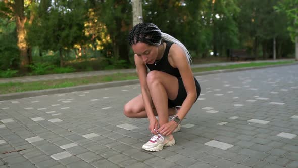 Stylish Woman Runner with Dreadlocks in Sport Wear Tying the Shoelaces on Pavement Road in the