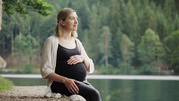 Pregnant woman touching gently her tummy when sitting near the mountain lake.