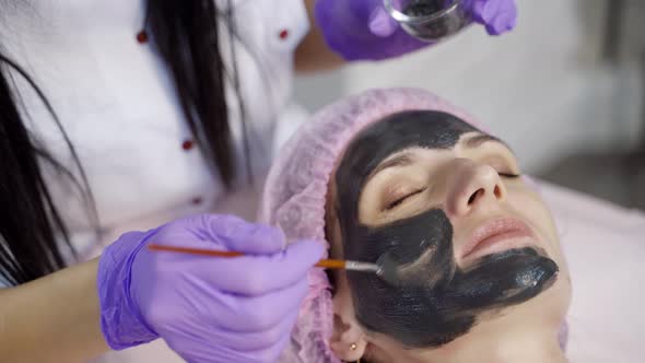 Cosmetologist Applies a Black Mask with a Brush to Cleanse and Moisturize the Skin of the Face