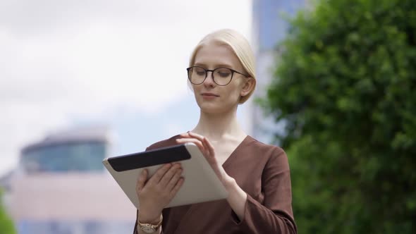 A Young Blonde Woman in Glasses Scrolls the Tablet with Interest in the Street