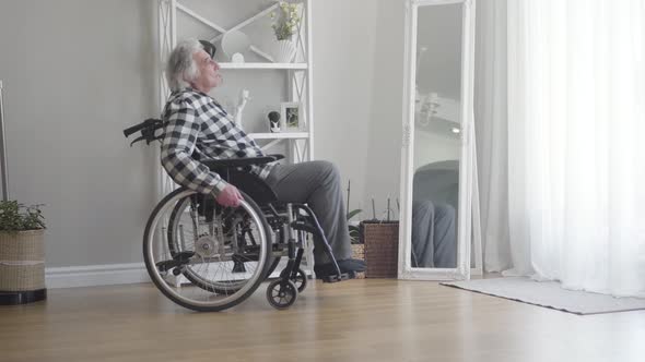 Side View of Upset Elderly Caucasian Man in Wheelchair Rolling To Window at Home. Depressed Old