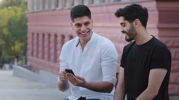 Couple of Young Attractive Indian Arab Guys Sitting Outdoors Holding Mobile Phone Look at Smartphone