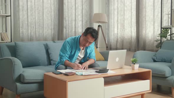 Asian Man With A Laptop Looking At The Bill And Recording The Expenses In Notebook