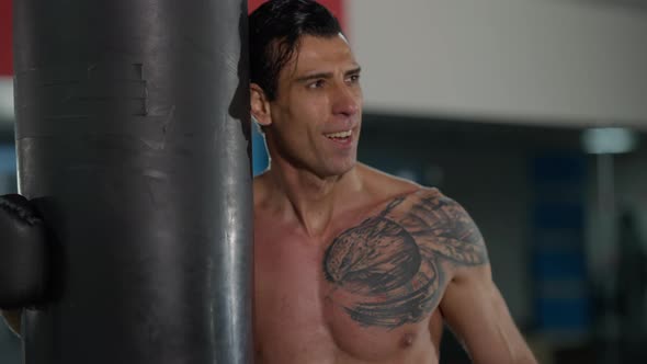 Tired Satisfied Perspiring Sportsman with Tattooed Chest Standing at Punching Bag Looking at Camera