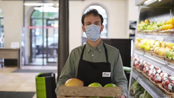 Portrait of Handsome Man in Face Mask and Apron Going Ahead in Food Store with Box of Fruits Indoors