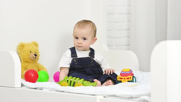 Baby Boy in Denim Overalls Sitting on the Bed Plays Toys and Throws Plastic Colored Balls