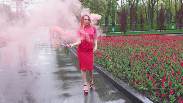 A Girl with Makeup with Rainbow Braids in Red Dress Posing in Red Smoke Against the Background of a