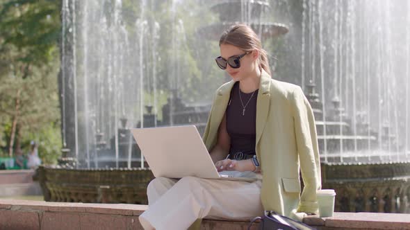 Elegant Young Woman Sitting on the Fountain in the City Center Working on the Computer