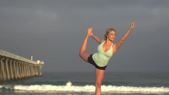 A young woman does yoga on the beach next to a pier.