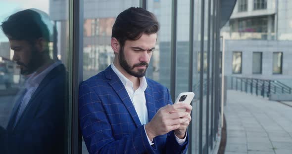 Male Millennial Professional Holding Modern Smartphone Texting Message Near the Glass Building