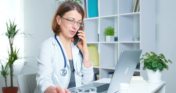 Portrait of a woman doctor consulting a patient on the phone and working with a laptop remotely. 4K.