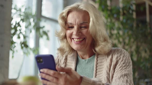 A Woman Aged Sitting at a Table and Watching a Video on the Phone