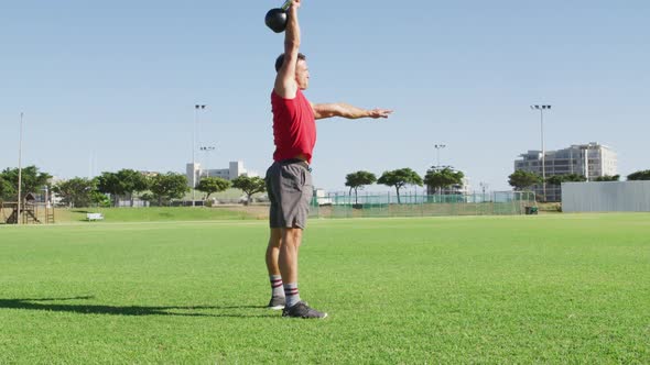 Fit caucasian man exercising outdoors, squatting and lifting kettlebell weight with one arm