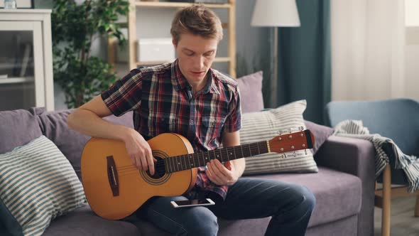 Young Musician Is Tuning Acoustic Guitar Touching Strings Sitting on Couch at Home During Leisure