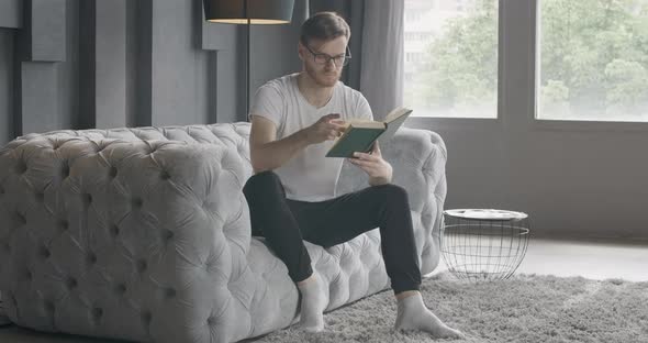 Wide Shot of Man Turning Pages of Book As Sitting on Couch Indoors. Portrait of Absorbed Young