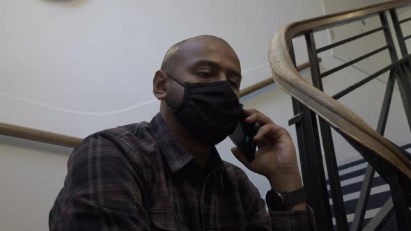 Adult Male Wearing Face Mask Using Mobile Phone Sitting Down On Stairwell. Locked Off, Low Angle