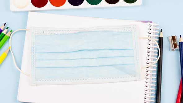 School Supplies Lie on a Pastel Blue Background with a Medical Mask