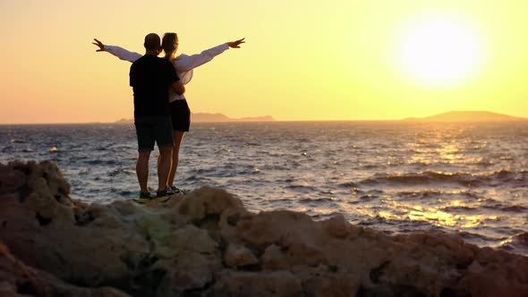 Loving Couple of Man and Woman Stands on Rock Against Backdrop of Sea and Setting Sun