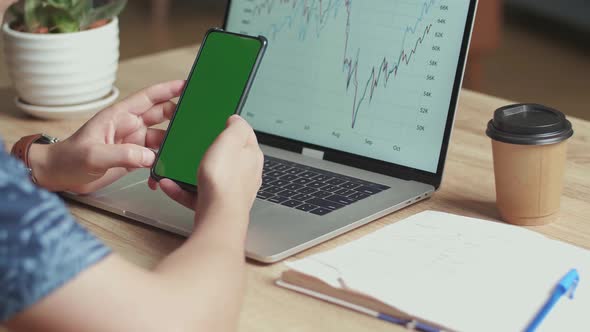 Man Use Mobile Phone With Green Screen Display, Financial Data On A Laptop Computer