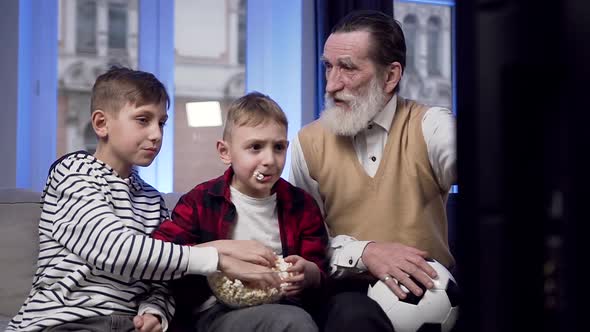 Boys which Eating Popcorn and Discussing with their Senior Bearded Granddad the Sport Game 