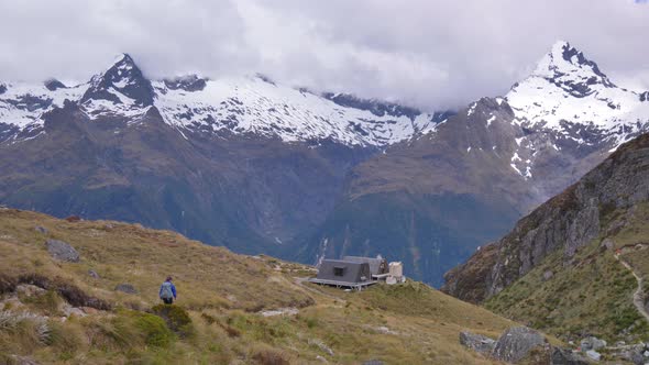 Pan, hiker walks towards remote alpine shelter, distant snow capped mountains, Routeburn Track New Z