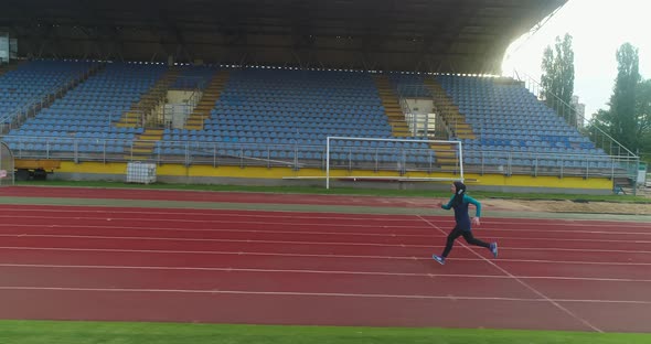 Side Aerial Tracking Shot of Muslim Woman on Soccer Stadium Exercising or Running Fast Wearing
