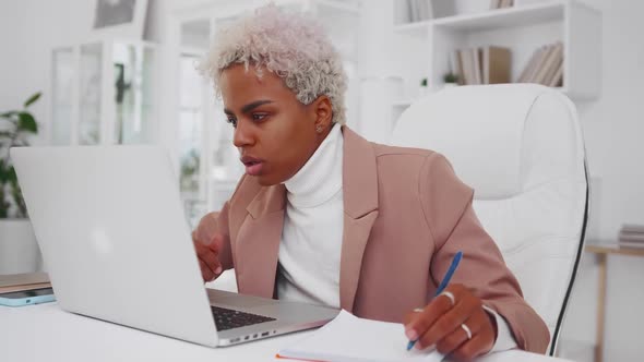 Young African American Woman is Writing Ideas in Notebook Looking at Laptop