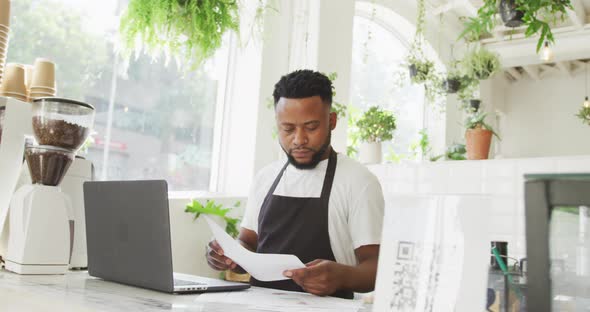 African american male cafe owner using laptop and looking at paperwork behind counter at cafe